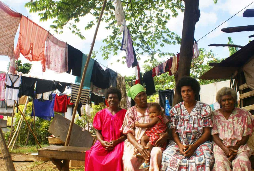 Mulheres e crianças sentadas à sombra, com roupa estendida a secar.