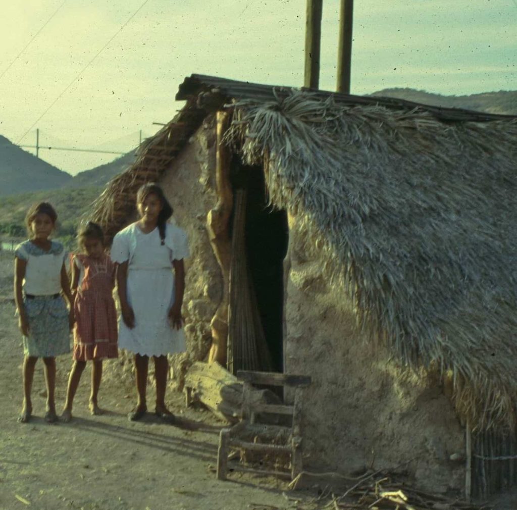 Três meninas diante de uma casa simples de palha.