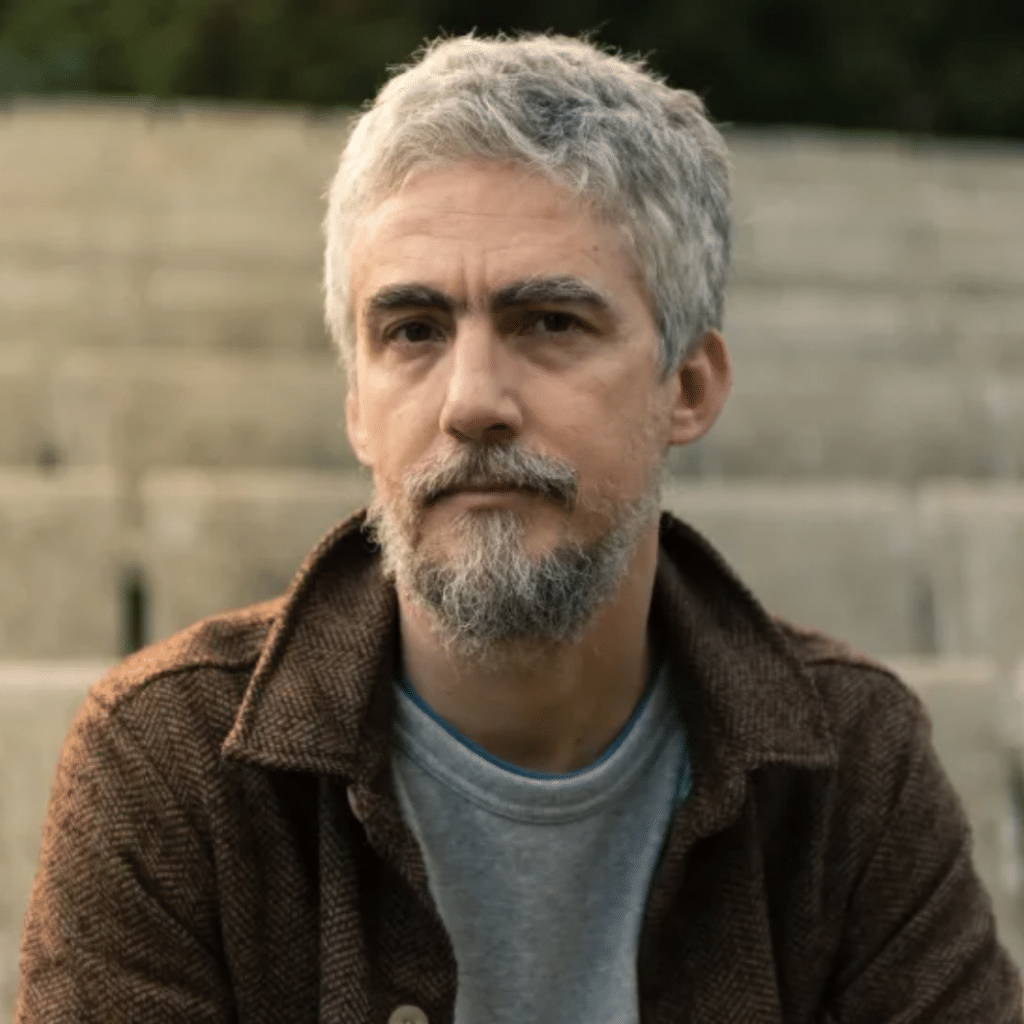 Homem com cabelo grisalho e barba, sentado em escadas.