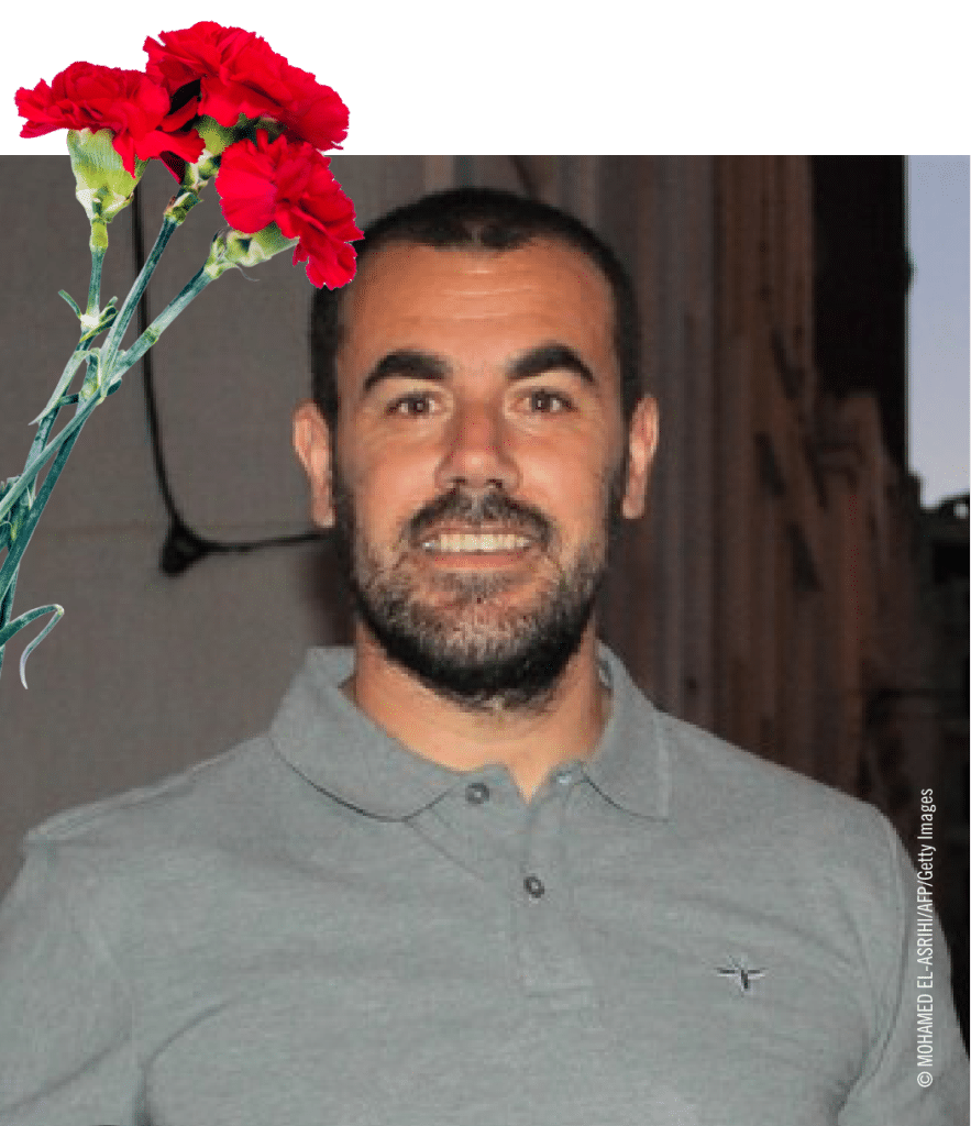 Homem sorridente com flores vermelhas ao lado.