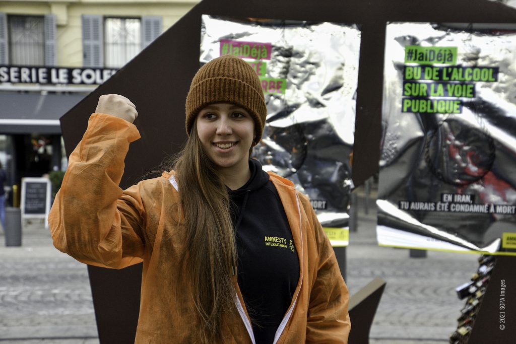 Activista com um punho cerrado, vestida de laranja, ao lado de cartazes sobre direitos humanos.