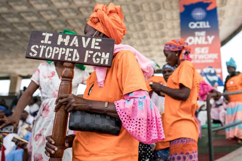 Uma mulher da Gâmbia, vestida de cor de laranja e cor-de-rosa, segura um cartaz onde se lê “Eu parei a MGF”. O Parlamento da Gâmbia rejeitou um projeto de lei para revogar a lei que proíbe a mutilação genital feminina. Foto © MUHAMADOU BITTAYE/AFP via Getty Images