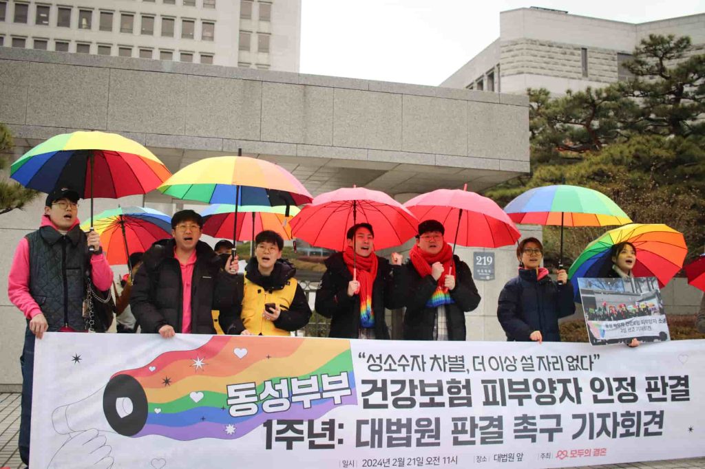 Sete pessoas da Coreia do Sul em fila, em frente a um cartaz, enquanto seguram guarda-chuvas coloridos. Foto © Amnistia Coreia do Sul