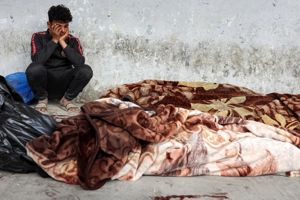 Um rapaz sentado ao lado dos corpos de vítimas mortais dos bombardeamentos israelitas em Gaza, embrulhados em cobertores no hospital Ahli Arab, também conhecido como hospital Batista, na Cidade de Gaza, em 18 de março de 2025. Foto de OMAR AL-QATTAA/AFP via Getty Images.