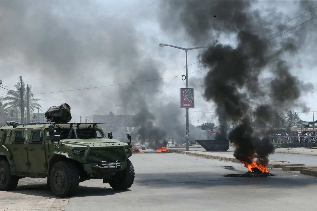 Veículo militar blindado junto a um pneu a arder durante uma manifestação contra o Governo em Maputo, a 6 de dezembro de 2024. Quase 300 pessoas foram mortas durante os confrontos entre manifestantes e forças de segurança desde as eleições moçambicanas de 9 de outubro de 2024. Foto de AMILTON NEVES/AFP via Getty Images.